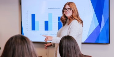 A woman gives a presentation in front of a screen with a chart. 