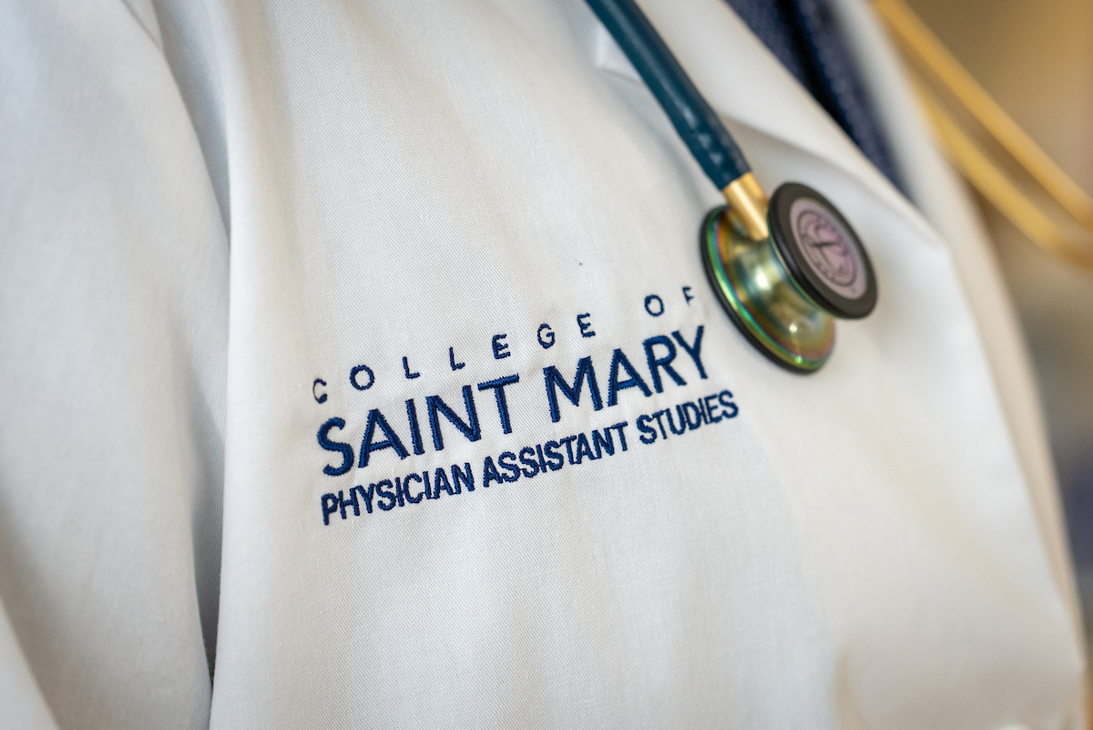 Detailed up close image of medical white coat with College of Saint Mary Physician Assistant Studies logo on it with the individual wearing a stethoscope.
