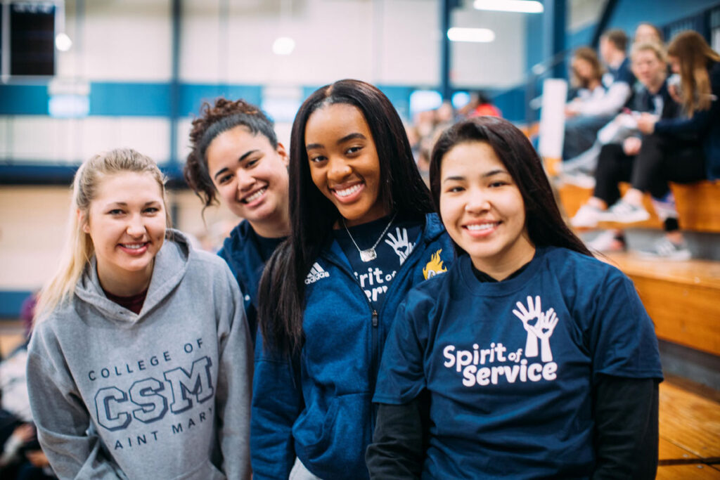 Four students in CSM and “Spirit of Service” shirts pose in a Lied Fitness Center Gymnasium at College of Saint Mary.