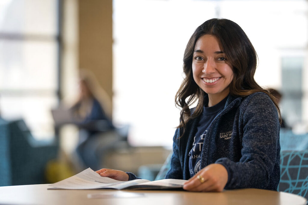 A student sits at a table reading an open book in a bright campus lounge.