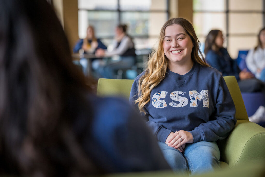 Student wearing a navy CSM sweatshirt sits in a campus lounge.
