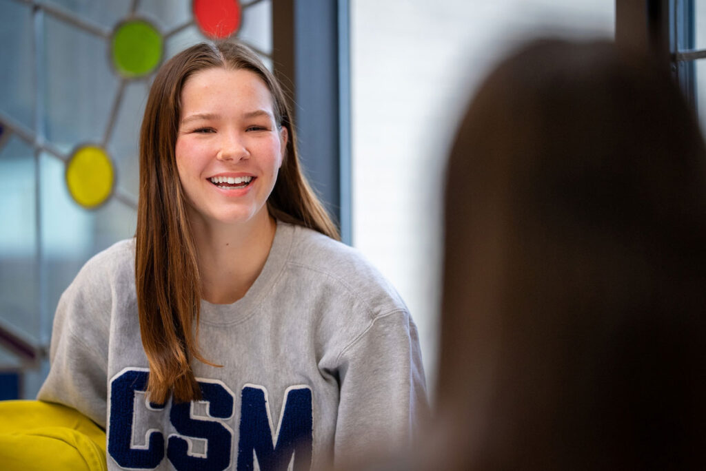 Student wearing a CSM sweatshirt sits in front of stained glass window in Hill-Macaluso Hall at College of Saint Mary.