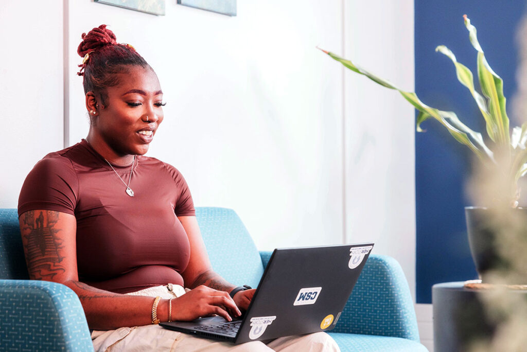 Student studies on a laptop with College of Saint Mary stickers on it while sitting in a campus lounge.