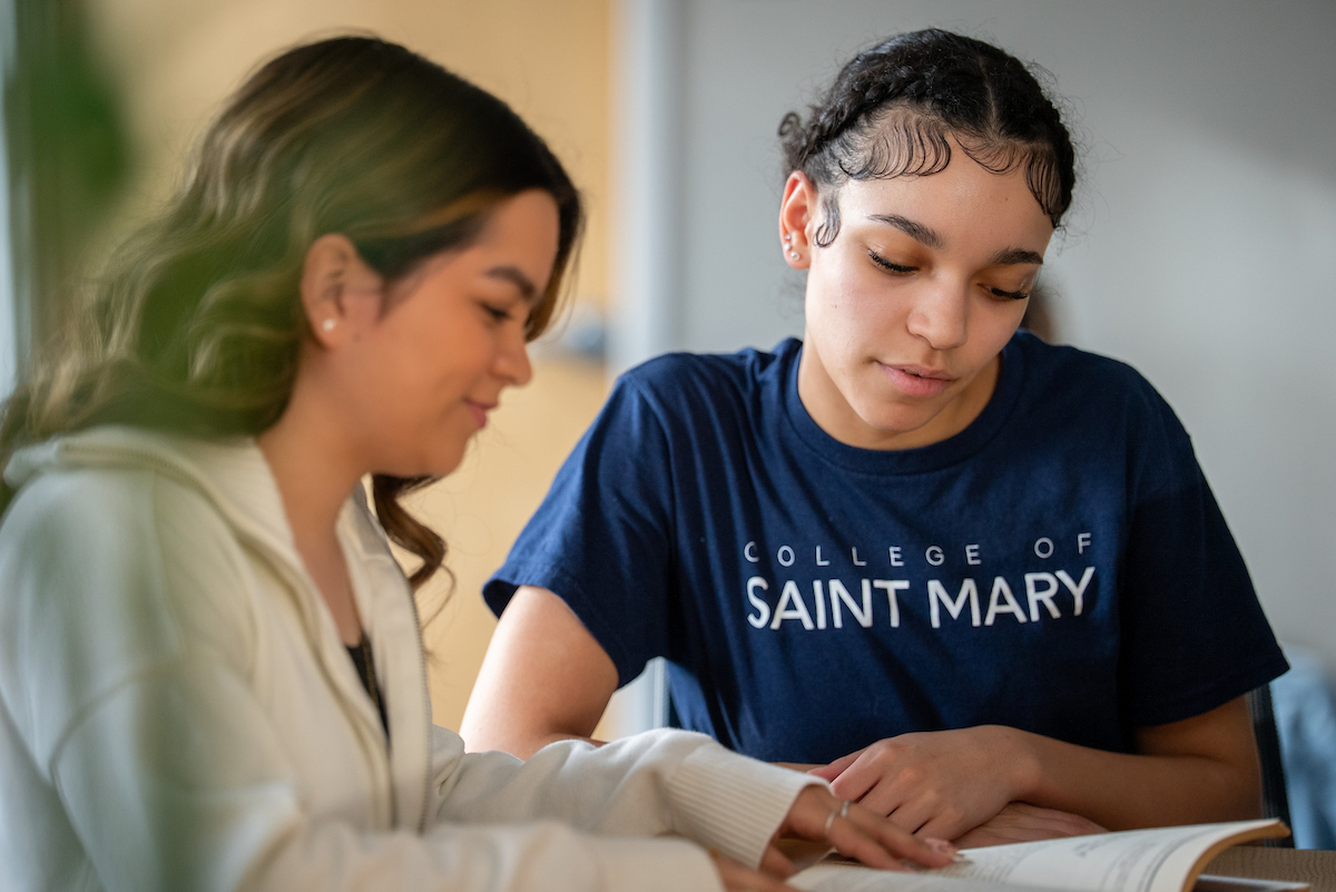 Two students discussing a book in a classroom, one wearing a College of Saint Mary T-shirt.