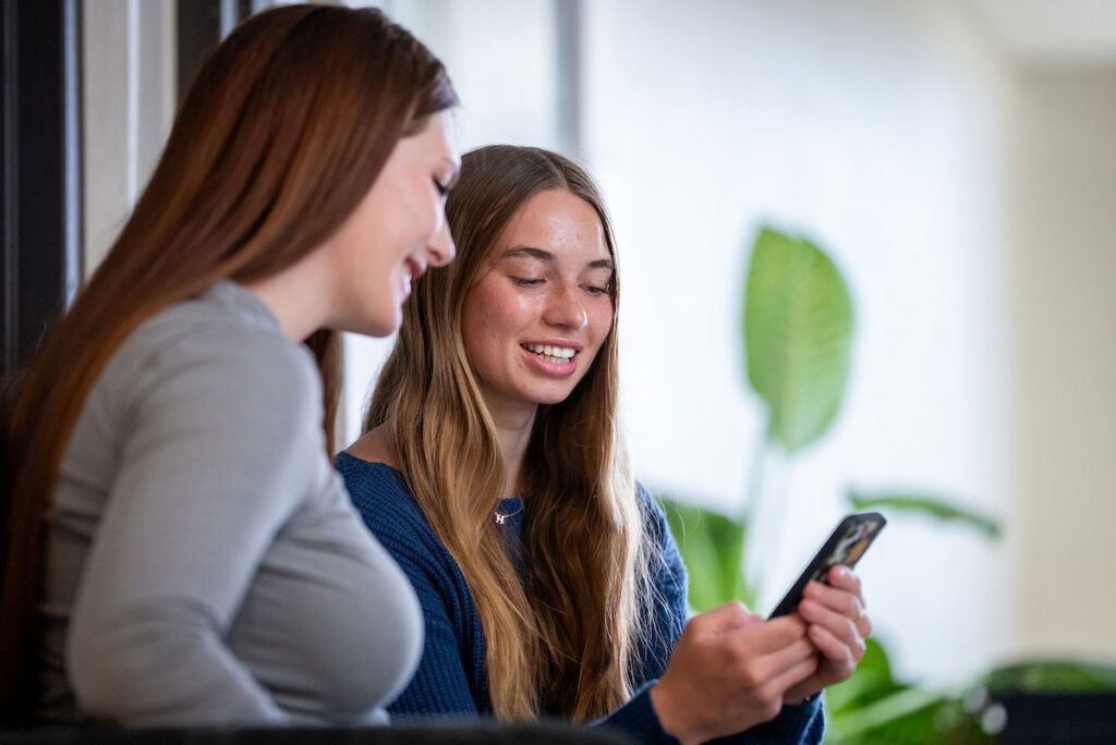 Two students scrolling on phone together and smiling.