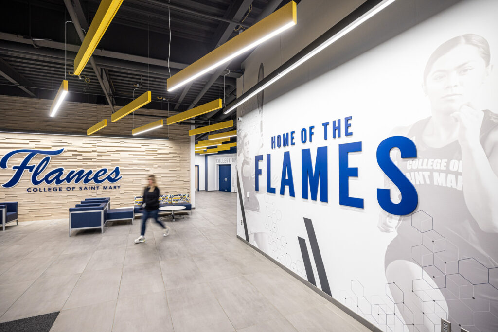 Wide hallway with a large wall graphic reading “Home of the Flames” and “Flames College Athletics,” with students walking through a modern campus space.