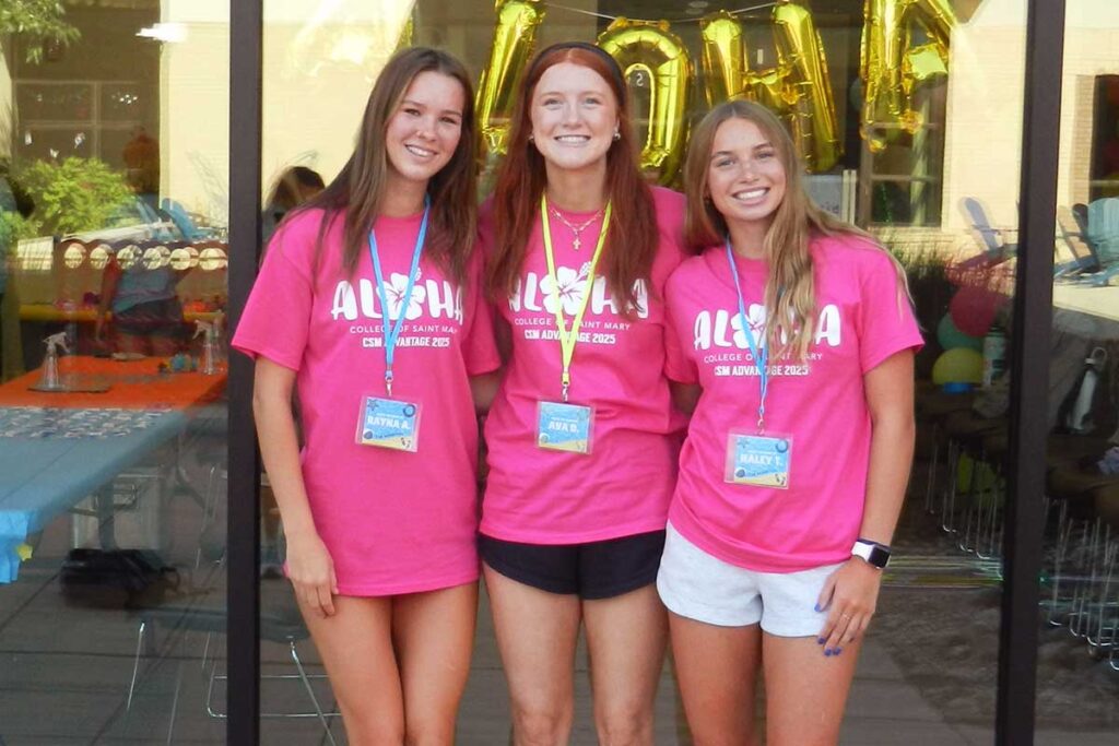 Three students wearing matching College of Saint Mary shirts stand together at a campus event, smiling in front of decorative balloons.