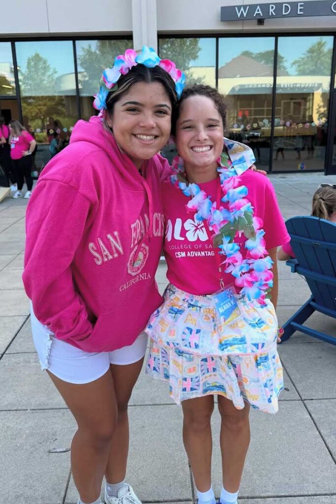 Two students wearing pink College of Saint Mary sweatshirts pose together outdoors during a campus event.
