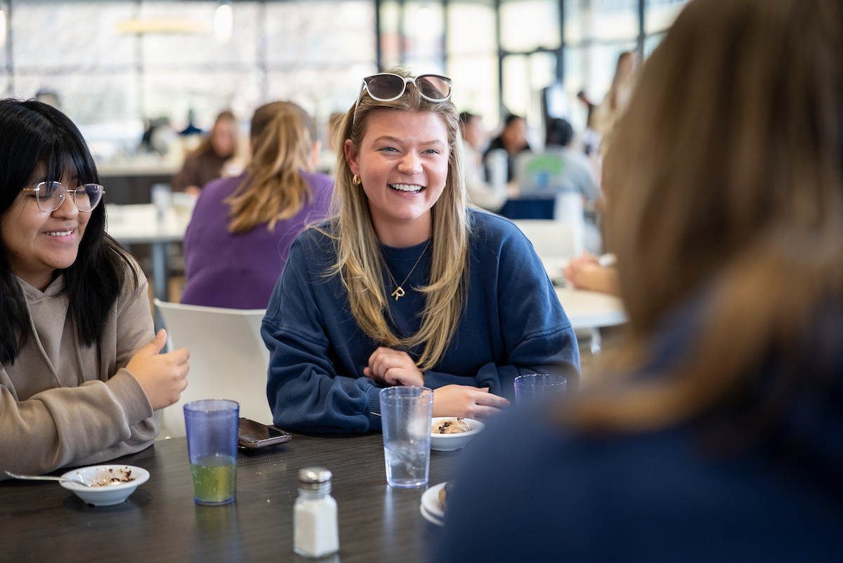 Students sitting together at a table in a campus dining area, talking and sharing a meal