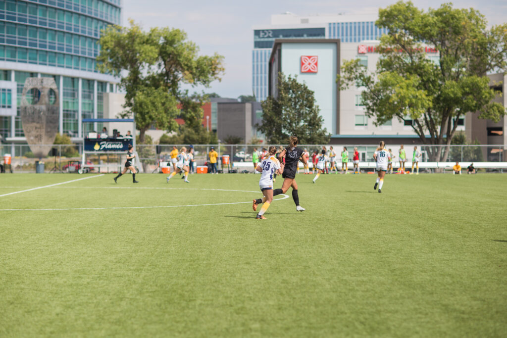 Students playing a soccer game on a green athletic field during a College of Saint Mary homecoming event, with campus buildings in the background.
