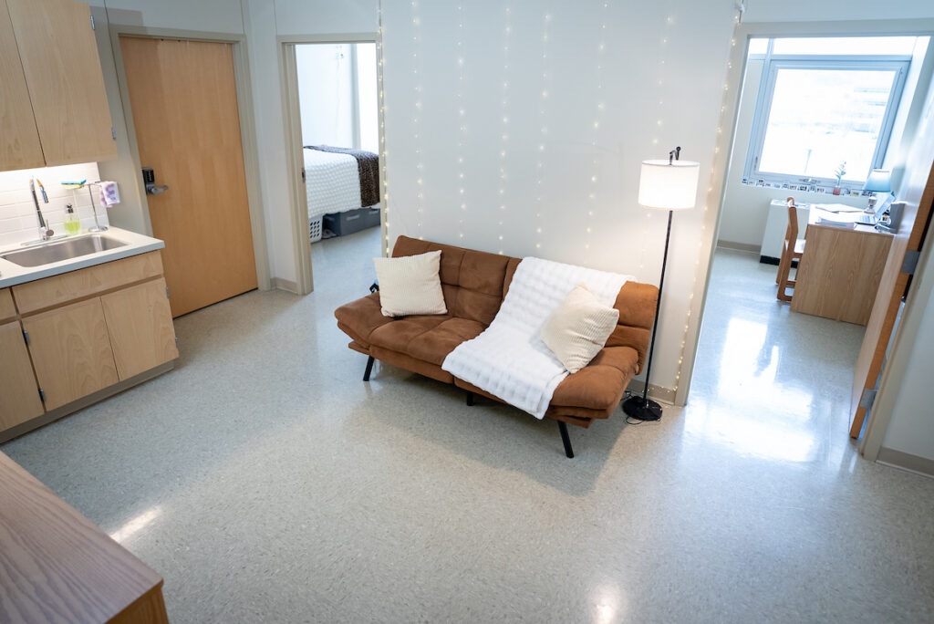Living and kitchenette area in Stevens Hall featuring a sofa, floor lamp, and kitchenette with cabinets in an apartment‑style residence hall.