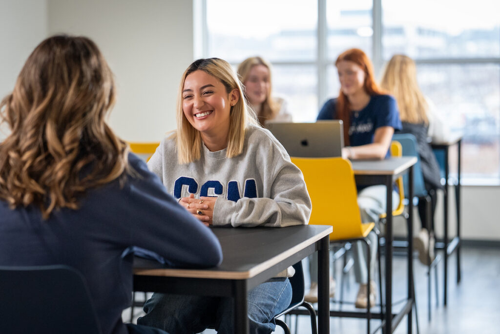 Students sitting at tables in a classroom or study space, working on laptops and talking together.