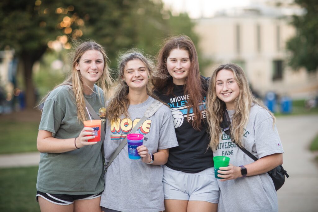 Four students standing together on campus, smiling and holding colorful cups during a campus life event.