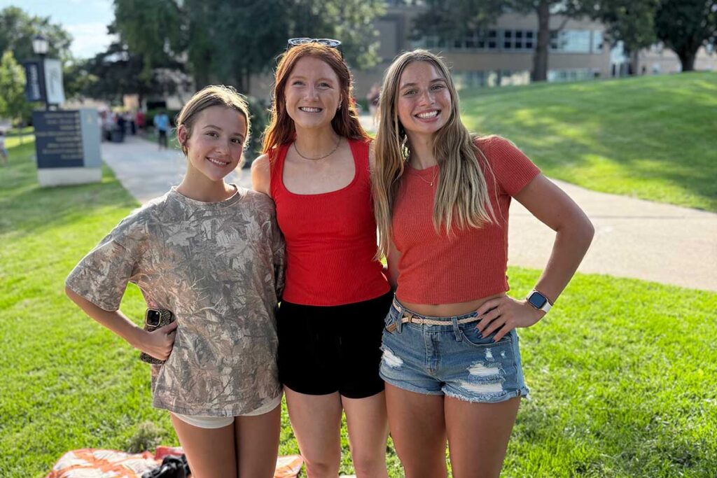 Three students standing together on campus, posing outdoors on a grassy lawn near campus buildings.