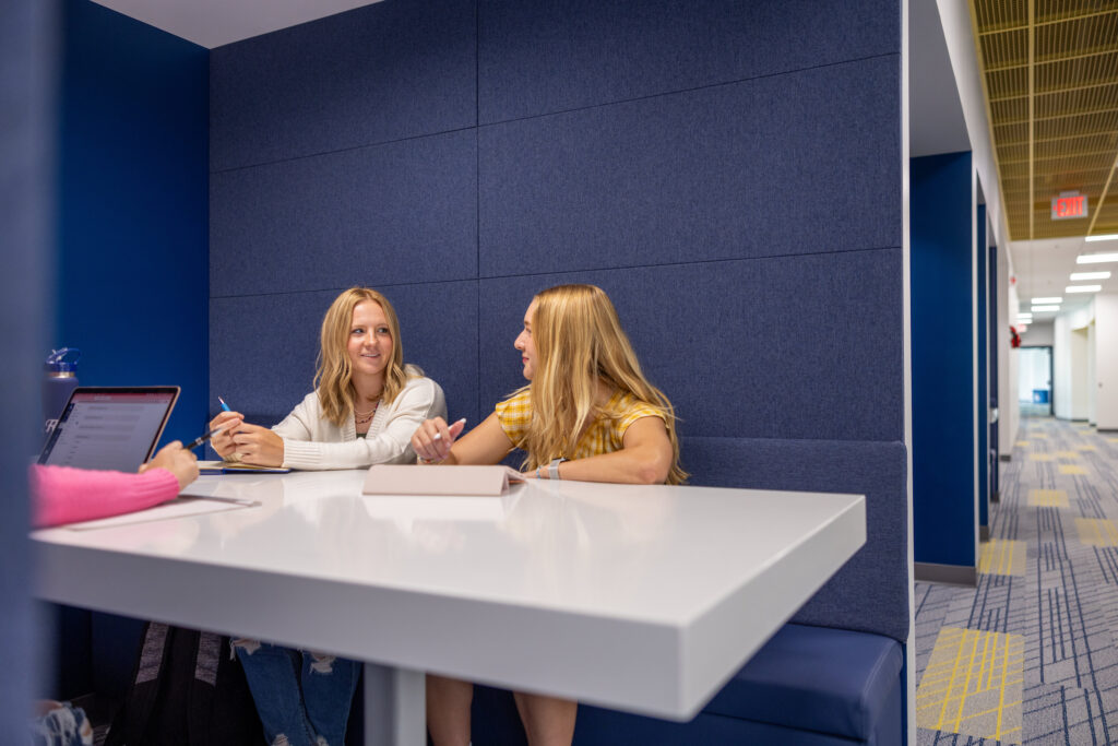 Interior photo of two students talking at a desk in front of blue wall
