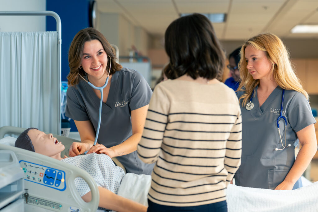 Two College of Saint Mary nursing students wearing grey scrubs practice their skills with a simulation patient with the guidance of a nursing professor.