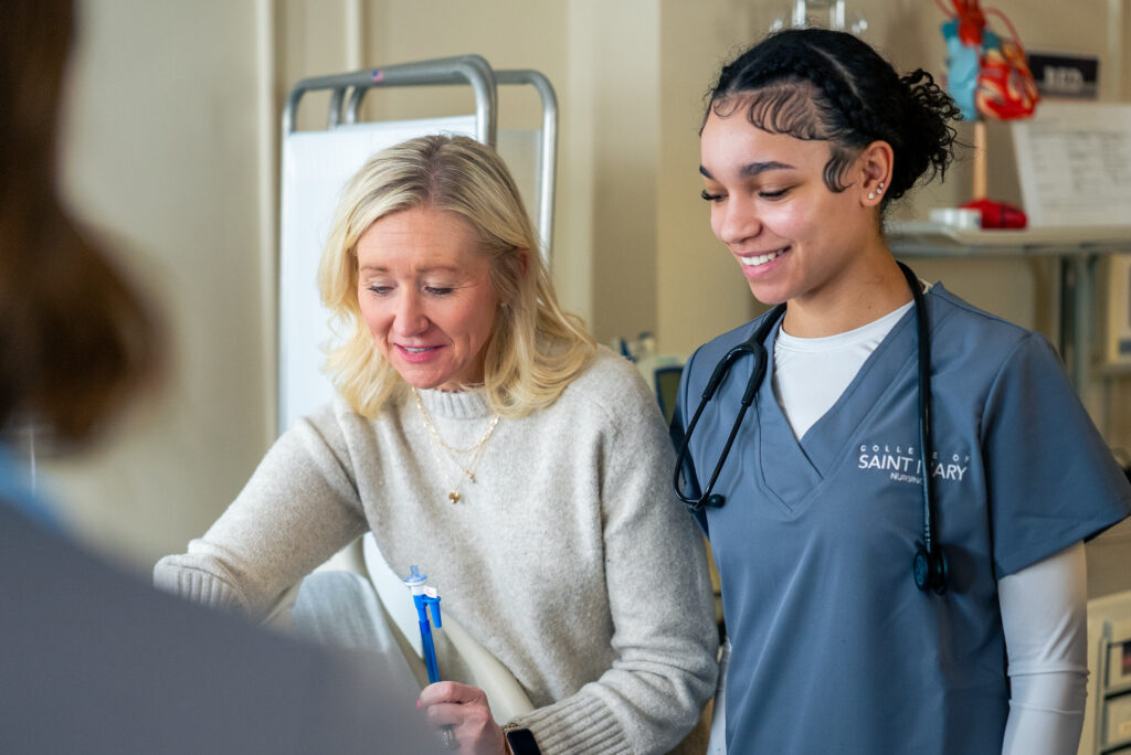 A nursing student wearing grey scrubs (right) learns from a nursing professor (left) in a lab.