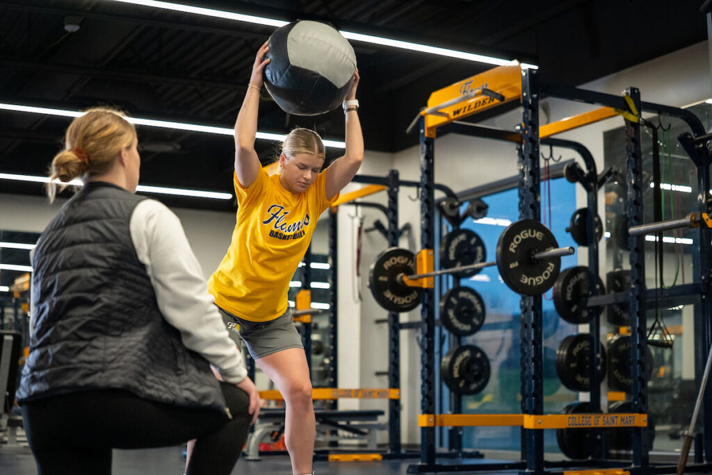 Student throws a medicine ball while instructor watches in a campus fitness center.
