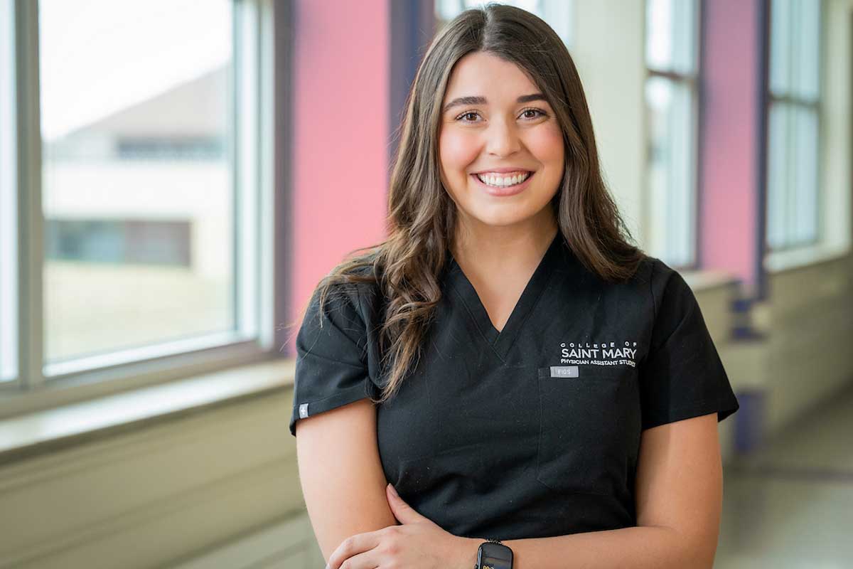 Physician Assistant student in black College of Saint Mary scrubs stands in power pose in classroom lab with bright natural lighting.