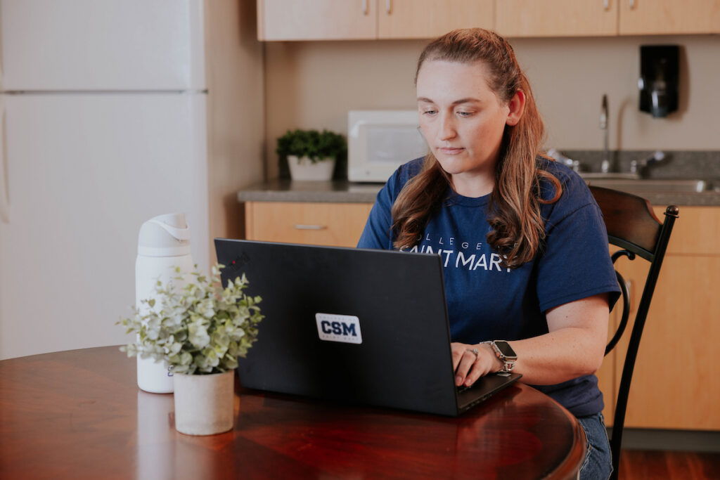 Student works on a laptop with College of Saint Mary sticker on it at a table.