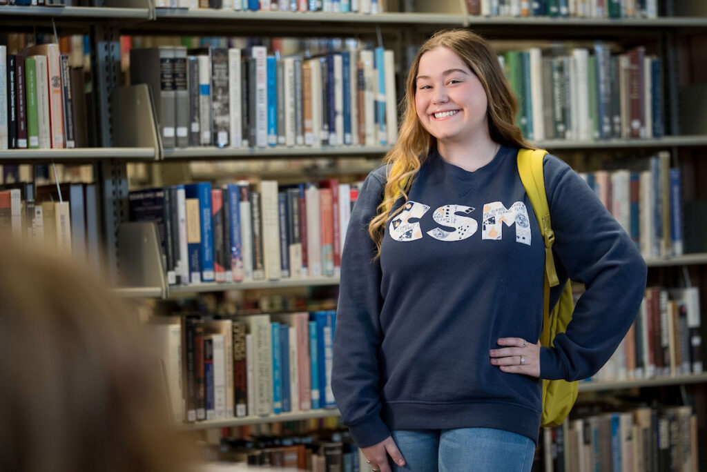 Student wearing a CSM navy sweatshirt stands in front of bookshelf in library at College of Saint Mary.