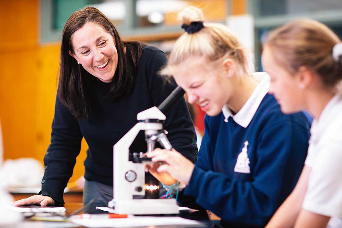 Instructor stands next to two students looking into microscope.