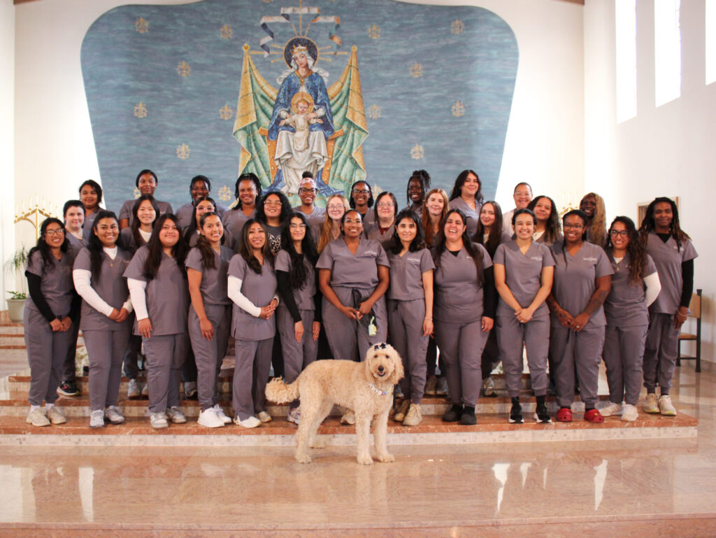 Practical nursing students wearing matching grey scrubs stand together in front of the religious mural in the CSM chapel. Stella, a golden doodle, stands in front of the group of students.