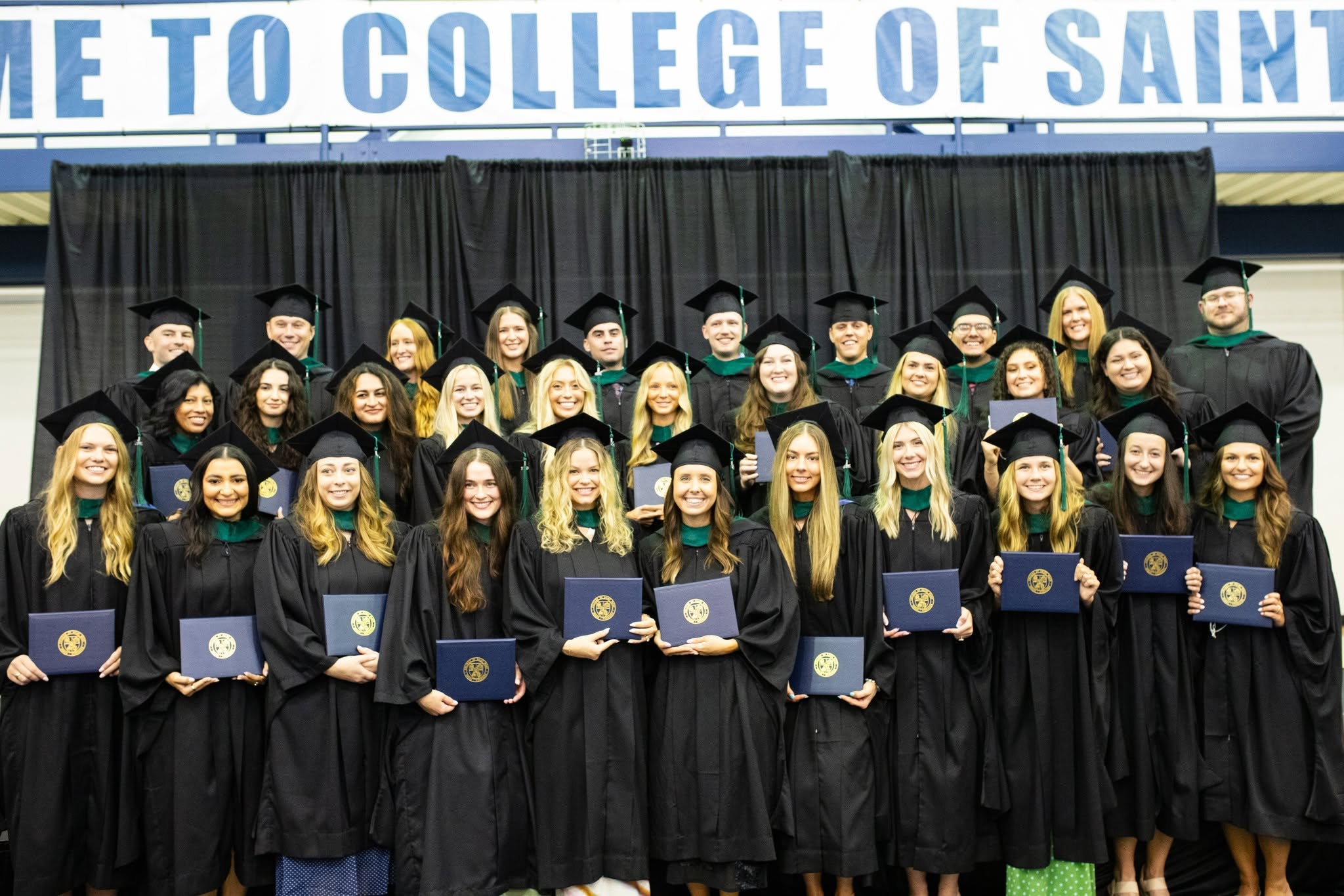 The physician assistant studies graduates stand together in a group wearing their black graduation caps and gowns holding their diplomas.