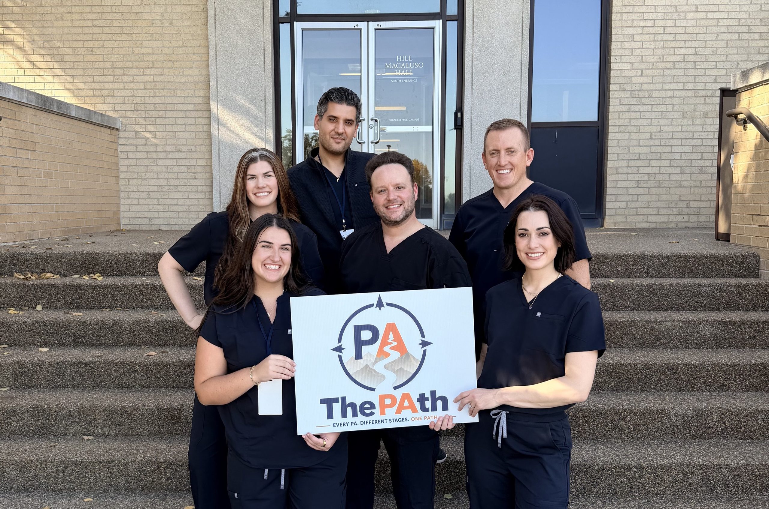 A group of practicing physician assistants wearing black scrubs stand in front of Hill Macaluso Hall at CSM holding a sign that says, "The PAth."